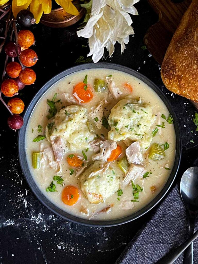 Homemade chicken and dumplings in a dark bowl with a loaf of bread in the background.