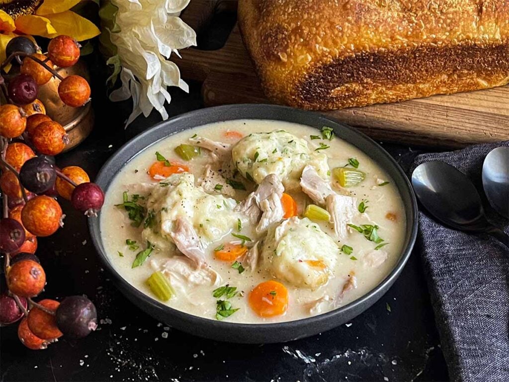 A bowl of chicken and dumplings with a loaf of bread in the background.