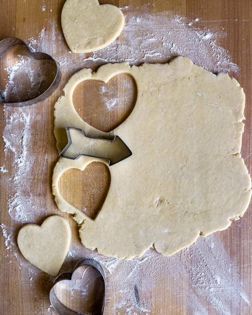 Soft frosted sugar cookie dough rolled out on a wooden board with heart shaped cookie cutters being used to cut out cookies.