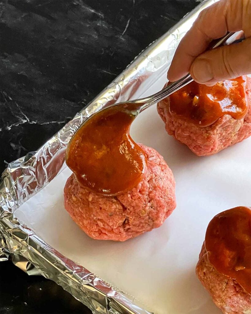 Unbaked mini meatloaves on a foil lined baking sheet with the sauce being spooned on top of them.