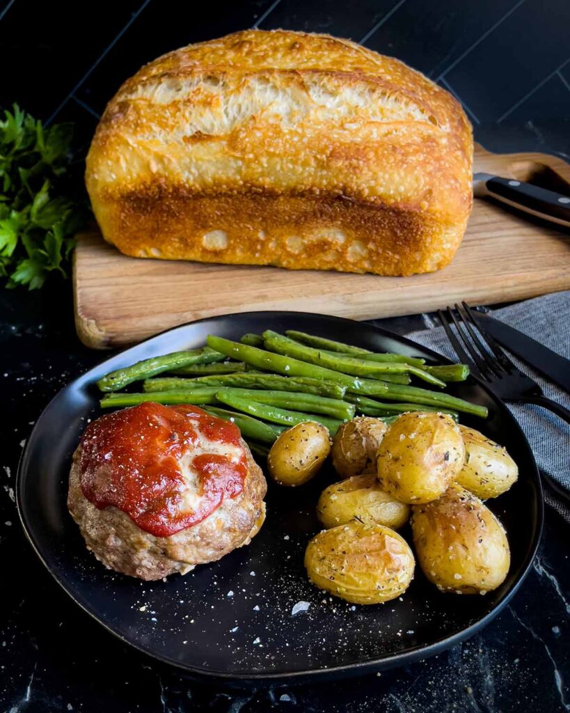 Mini meatloaf, roasted potatoes and green beans on a dark plate with a loaf of sourdough bread behind it.