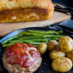 Mini meatloaf, roasted potatoes and green beans on a dark plate with a loaf of sourdough bread behind it.