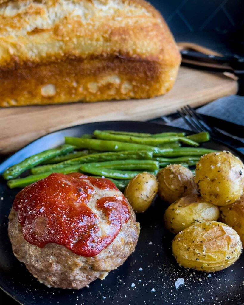Mini meatloaf, roasted potatoes and green beans on a dark plate with a loaf of sourdough bread behind it.