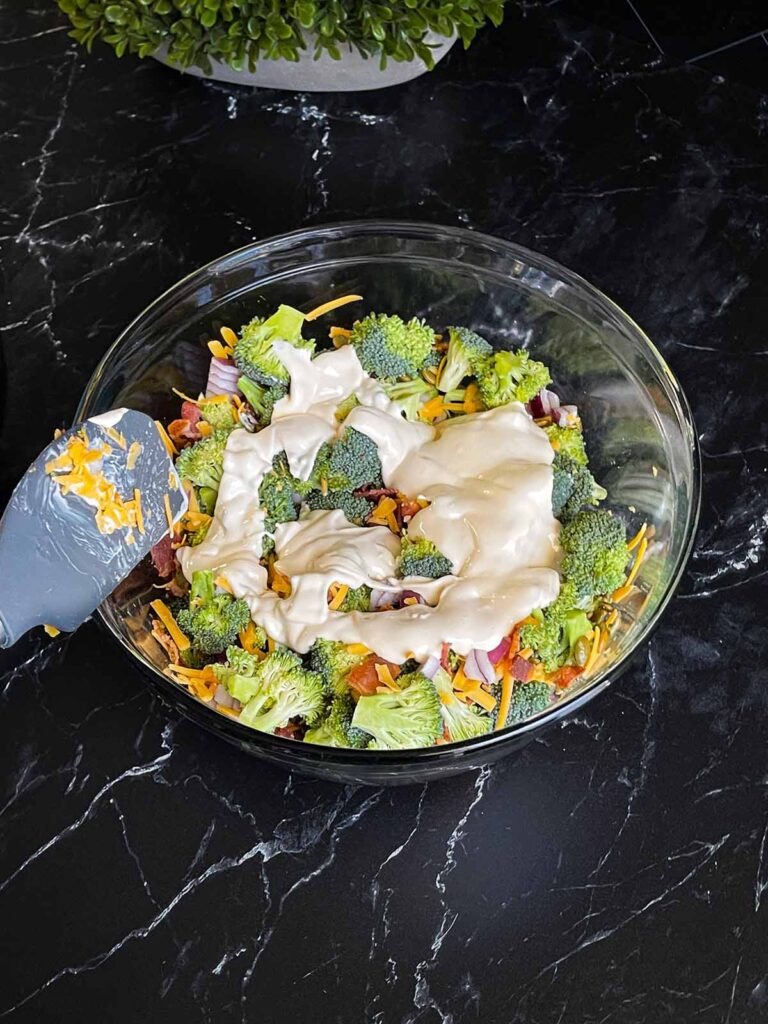 Dressing poured over the broccoli salad ingredients in a glass bowl.