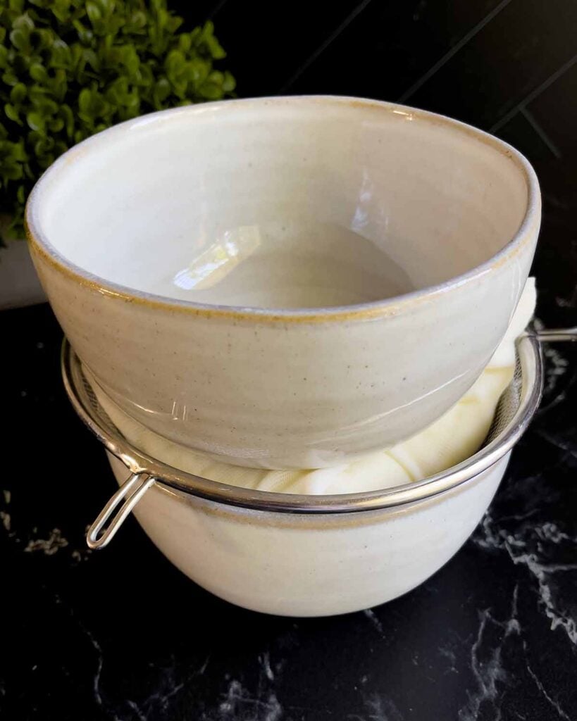 Ricotta cheese in cheesecloth and a strainer placed over a bowl, with another bowl weighing it down.