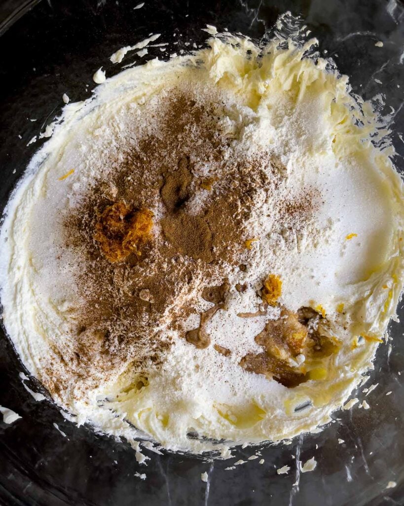 Granulated sugar, cinnamon, flour, orange zest, vanilla, and salt added to the cannoli cheesecake mixture in a glass bowl.