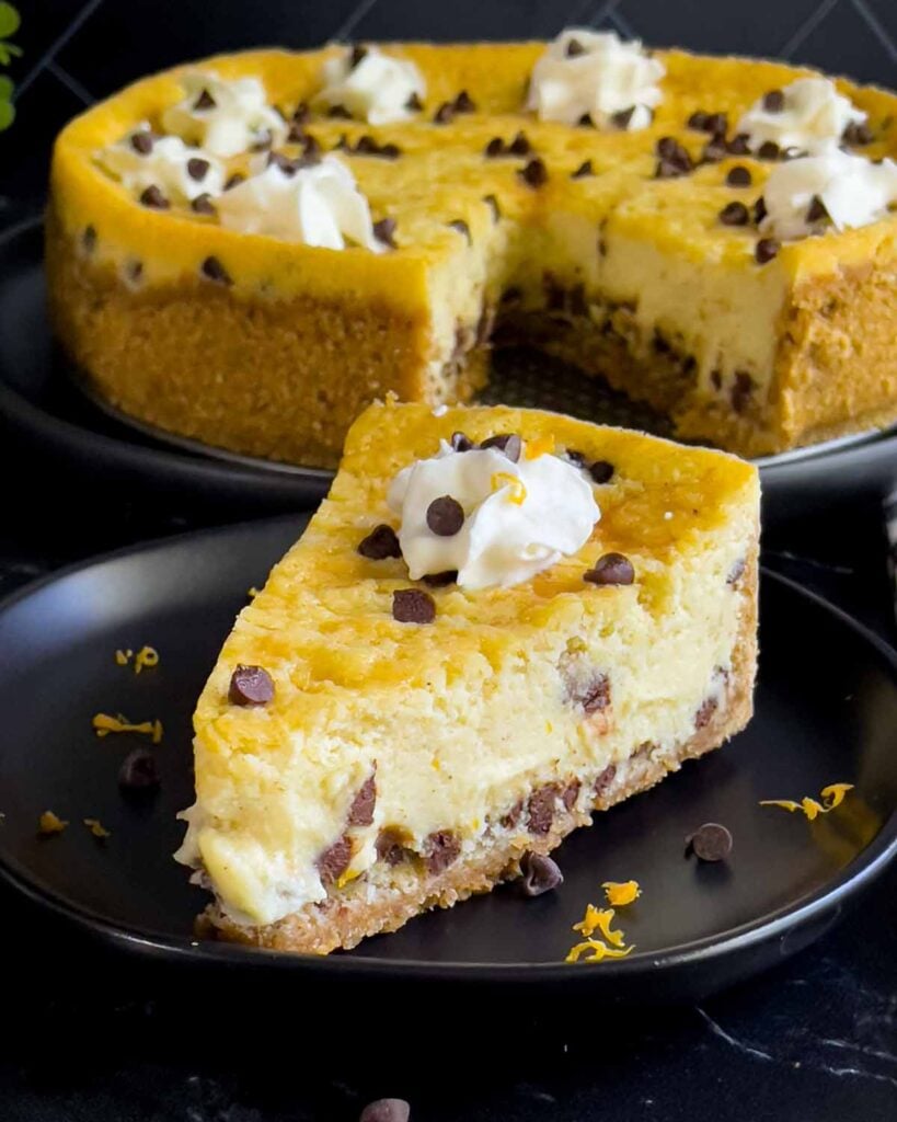 A slice of cannoli cheesecake on a dark plate with the whole cheesecake behind it.