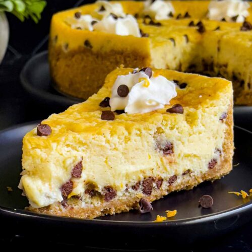 A slice of cannoli cheesecake on a dark plate with the whole cheesecake behind it.