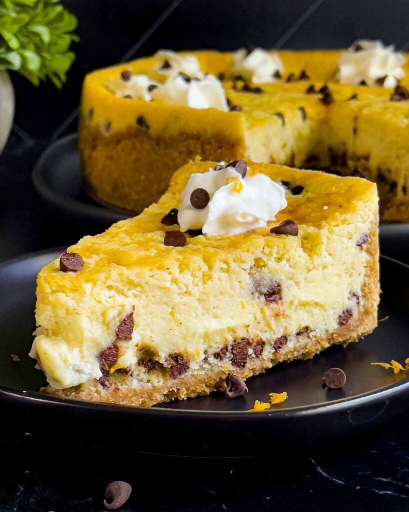 A slice of cannoli cheesecake on a dark plate with the whole cheesecake behind it.