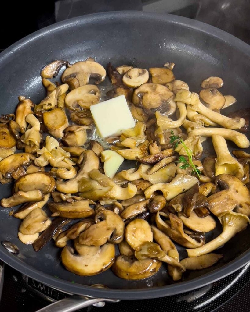 Mushrooms cooking in a skillet. Butter, fresh thyme, and a garlic clove added.