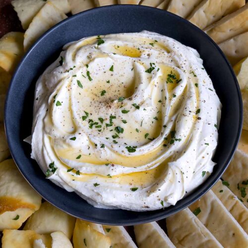 Whipped feta garnished with honey, chopped parsley, and black pepper in a dark bowl surrounded by pita chips and pita bread.