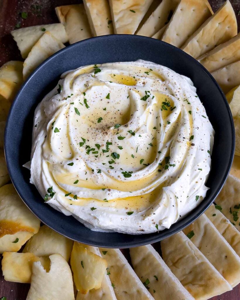 Whipped feta garnished with honey, chopped parsley, and black pepper in a dark bowl surrounded by pita chips and pita bread.