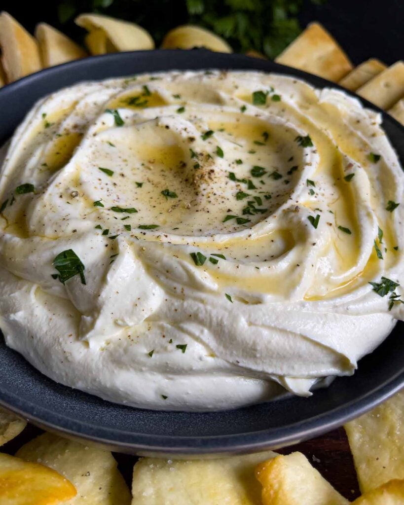 Whipped feta garnished with honey, chopped parsley, and black pepper in a dark bowl surrounded by pita chips and pita bread.
