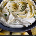 Whipped feta garnished with honey, chopped parsley, and black pepper in a dark bowl surrounded by pita chips and pita bread.