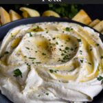 Whipped feta garnished with honey, chopped parsley, and black pepper in a dark bowl surrounded by pita chips and pita bread.