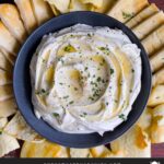 Whipped feta garnished with honey, chopped parsley, and black pepper in a dark bowl surrounded by pita chips and pita bread.