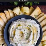 Whipped feta garnished with honey, chopped parsley, and black pepper in a dark bowl surrounded by pita chips and pita bread.