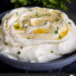 Whipped feta garnished with honey, chopped parsley, and black pepper in a dark bowl surrounded by pita chips and pita bread.