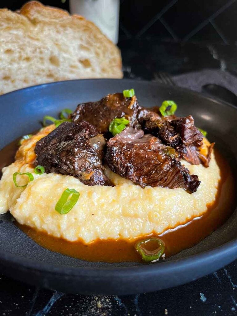 Braised beef cheek on a bed of polenta in a dark bowl.