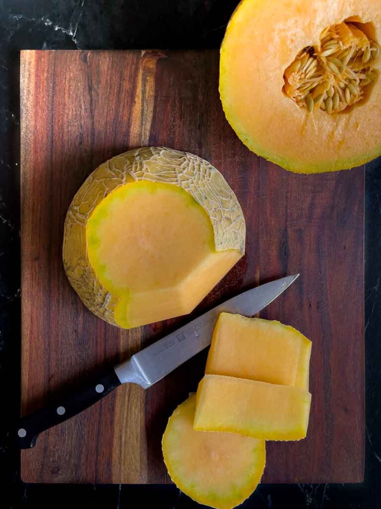 Cantaloupe cut in half and skin cut off on a dark cutting board.