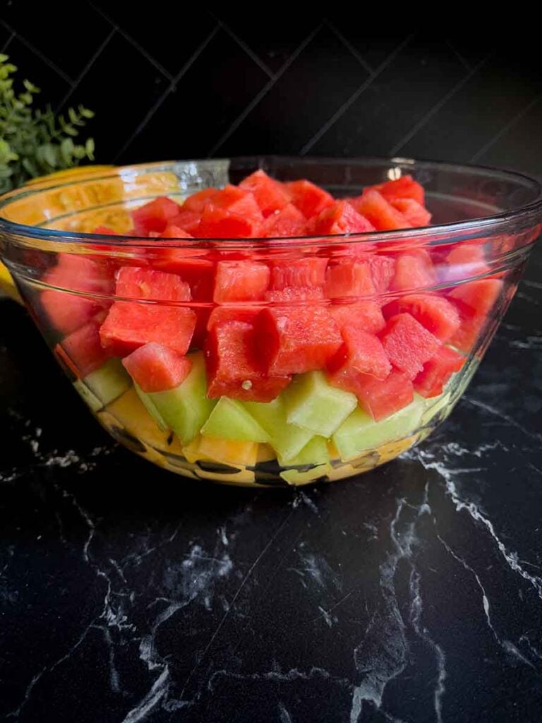 Watermelon cubes, honeydew melon cubes, and cantaloupe cubes in a glass mixing bowl on a dark surface.