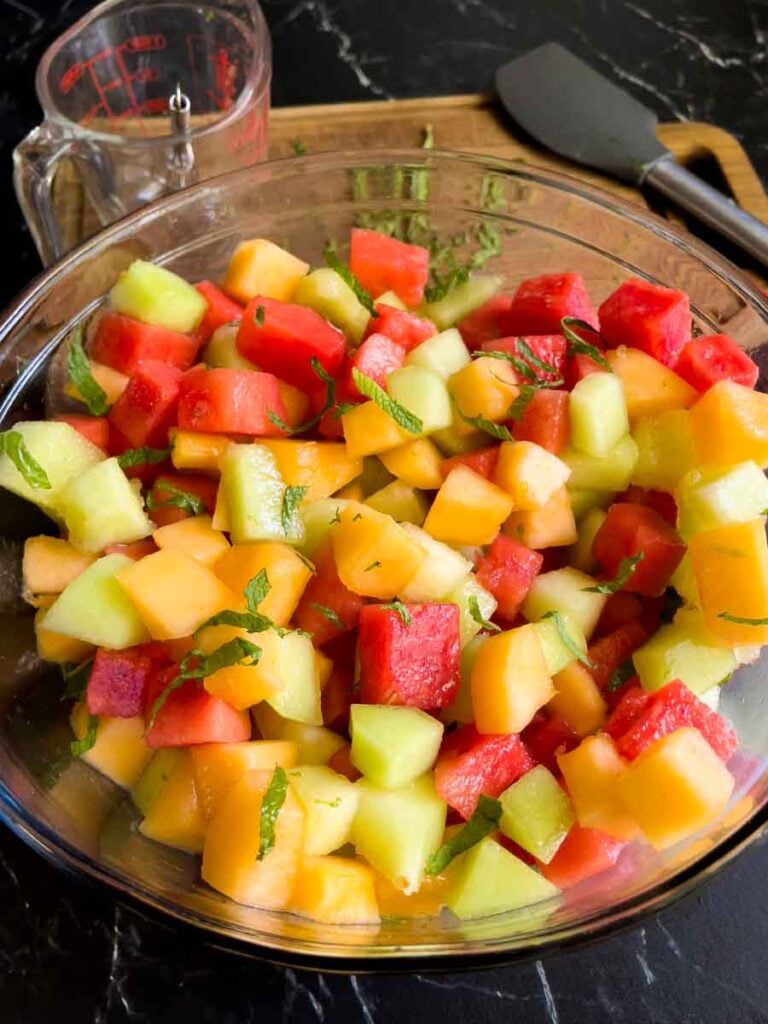 Dressed melon fruit salad in a large glass mixing bowl on a dark surface.