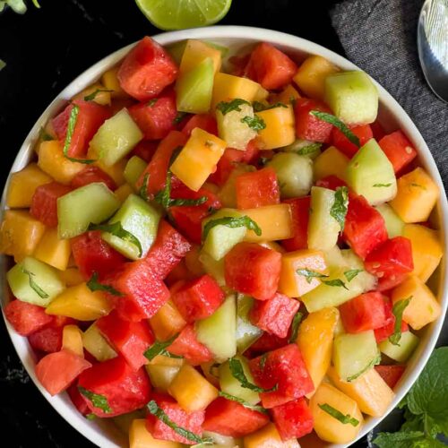 Mixed melon fruit salad in a white bowl on a dark surface.