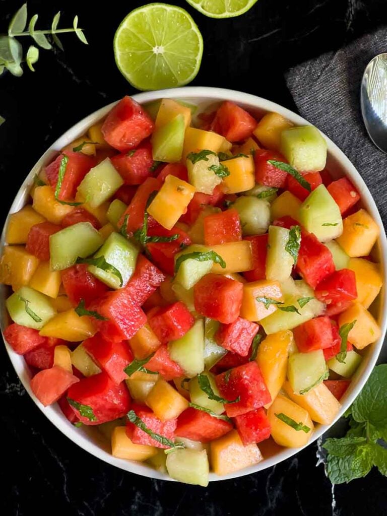 Mixed melon fruit salad in a white bowl on a dark surface.