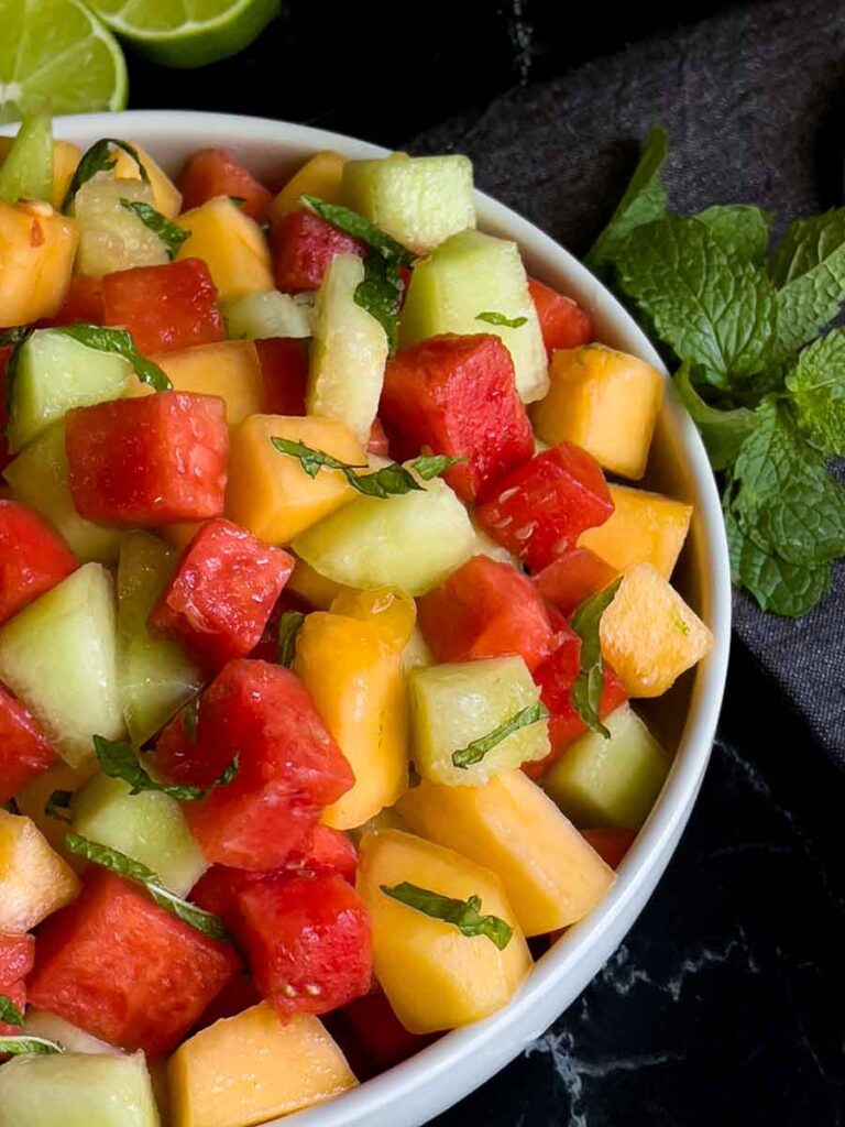 Mixed melon fruit salad in a white bowl on a dark surface.