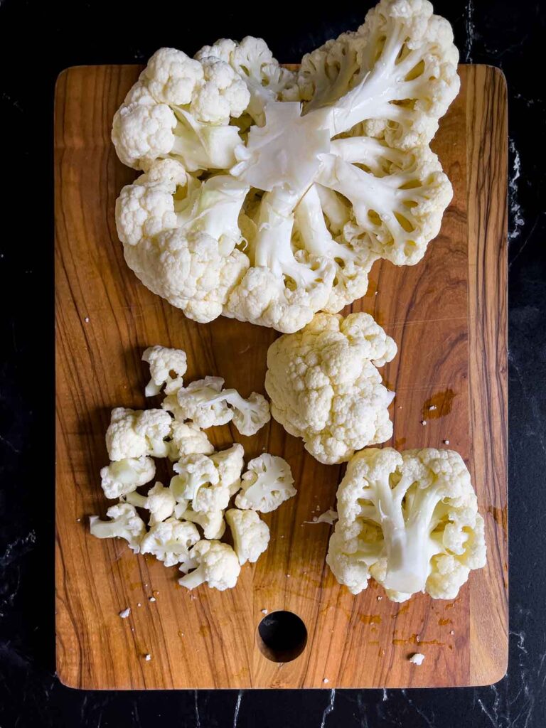 Head of cauliflower being cut into small pieces on a wooden cutting board.