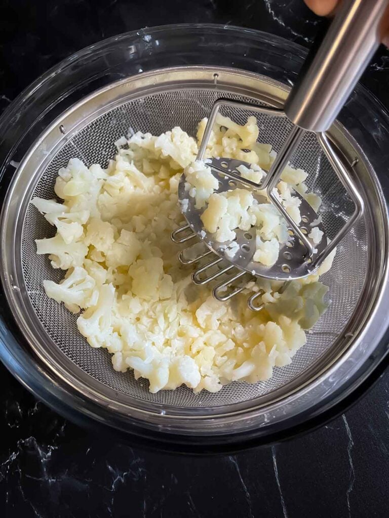 Cooked cauliflower in a fine mesh strainer being mashed with a potato masher.
