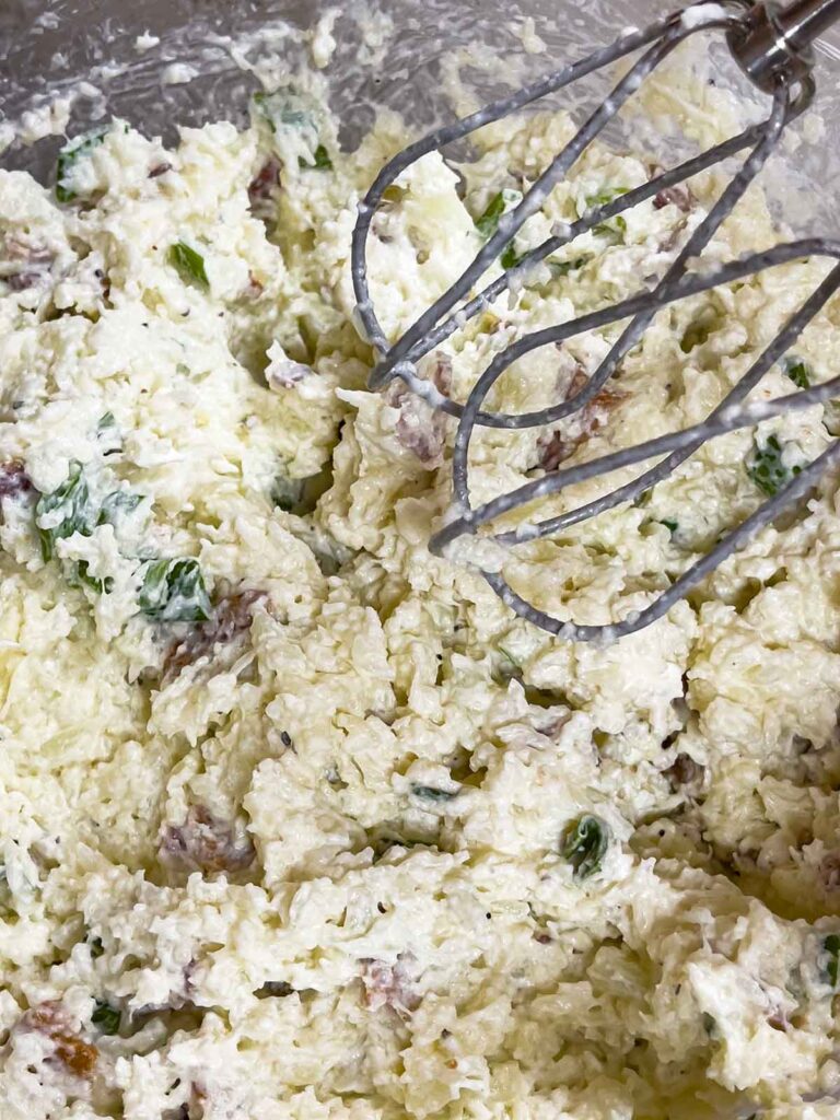 The loaded cauliflower mash in a glass bowl being mixed with an electric handheld mixer.