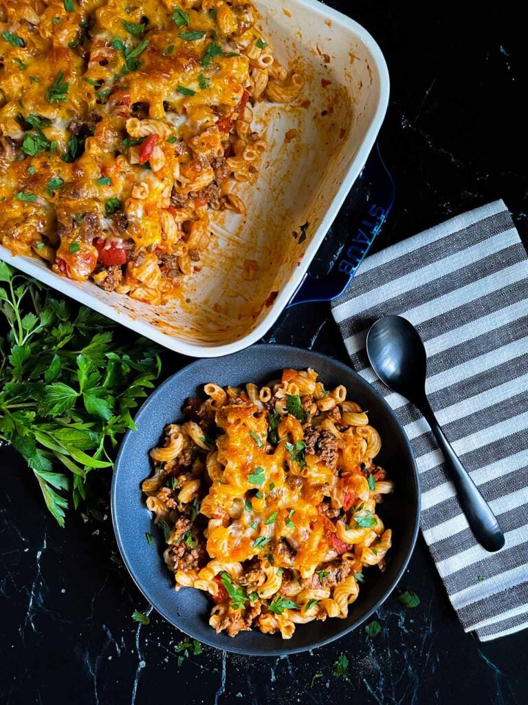 Cheesy beefaroni served in a dark bowl with the casserole off to the side.