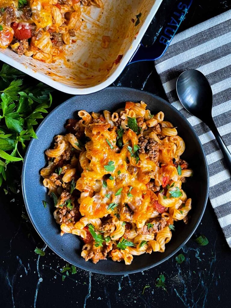 Cheesy beefaroni served in a dark bowl with the casserole off to the side.