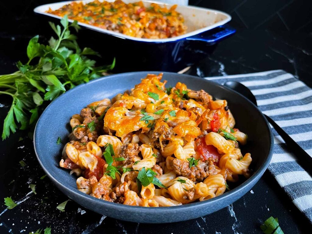 Cheesy beefaroni served in a dark bowl with the casserole off to the side.