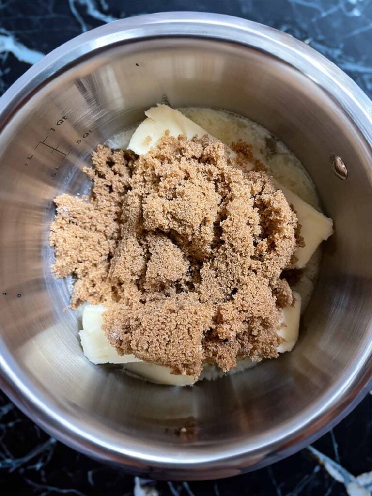 Dark brown sugar and butter for oyster cracker churro snack mix in a silver saucepan on a dark surface.