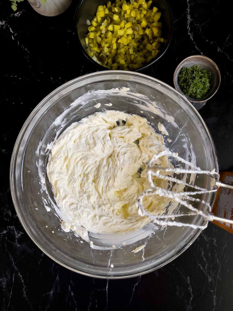 Blended cream cheese in a glass bowl with dill pickles and dill in ramekins on a dark surface.