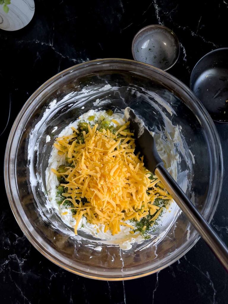 Cheddar cheese, dill, and diced dill pickles added to the cream cheese mixture in a glass bowl on a dark surface.