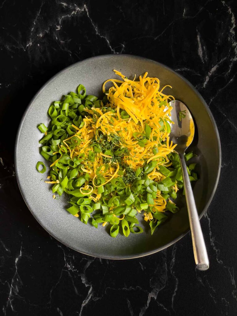 Green onions, dill, and cheddar cheese toppings for the dill pickle cheese ball in a dark bowl on a dark surface.