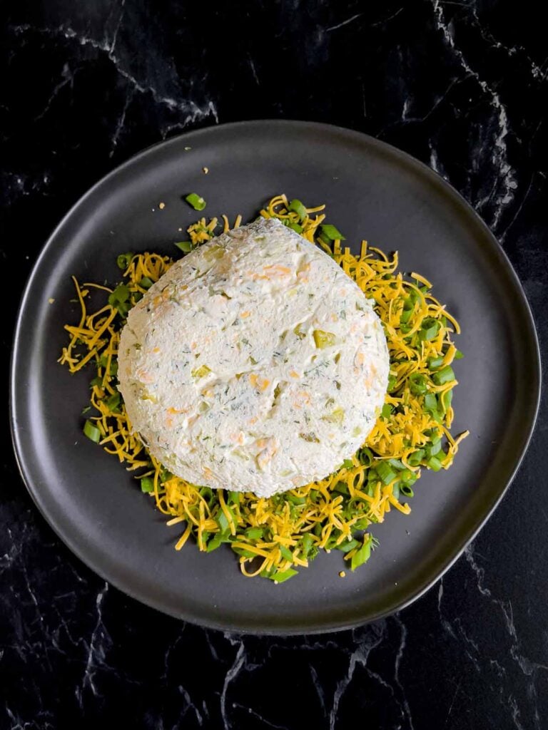 Dill pickle cheese ball on a bed of garnish of cheddar cheese, dill, and green onions on a dark plate on a dark surface.