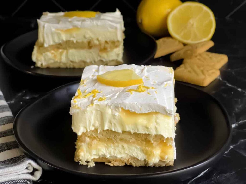 A serving of Lemon Icebox cake garnished with a lemon slice and zest on a dark plate.