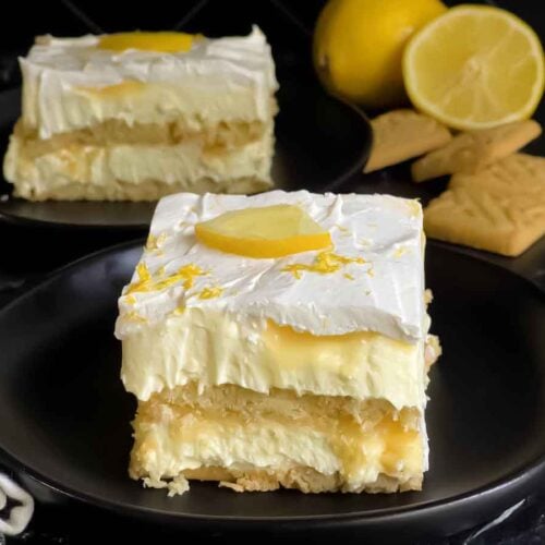 A serving of Lemon Icebox cake garnished with a lemon slice and zest on a dark plate.