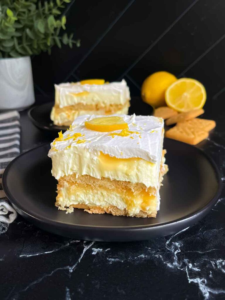 A serving of Lemon Icebox cake garnished with a lemon slice and zest on a dark plate.