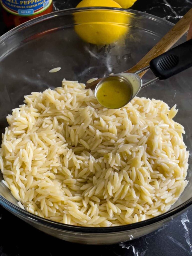 Drained orzo in a glass bowl tossing with a small amount of dressing and olive oil.