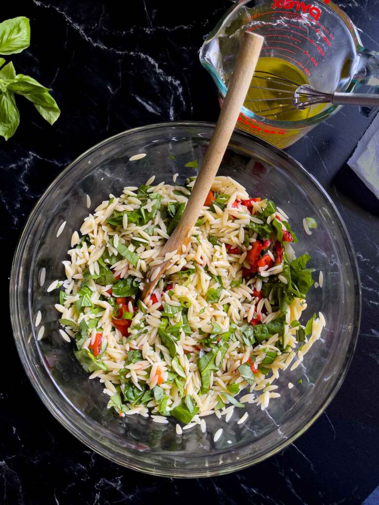 Vegetables added to the orzo in a glass bowl with orzo salad dressing in a glass measuring cup.