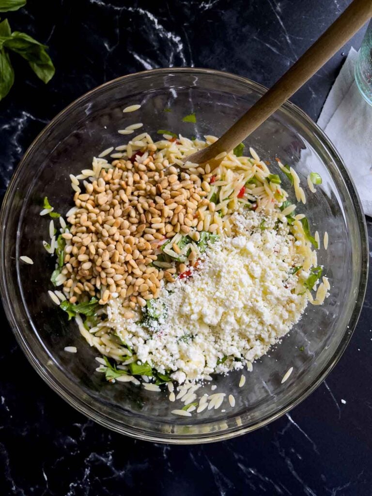 Toasted pine nuts and crumbled feta cheese added to the spinach and feta orzo salad in a glass bowl on a dark surface.