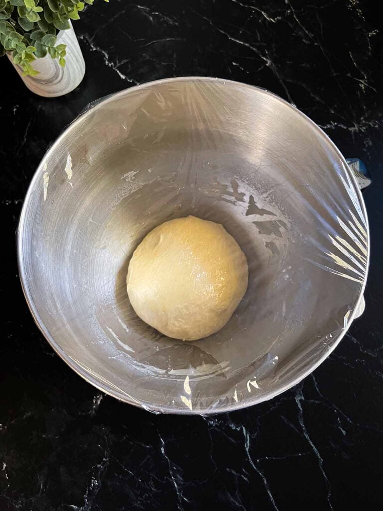 Pizza dough covered with plastic wrap in a stand mixer bowl.