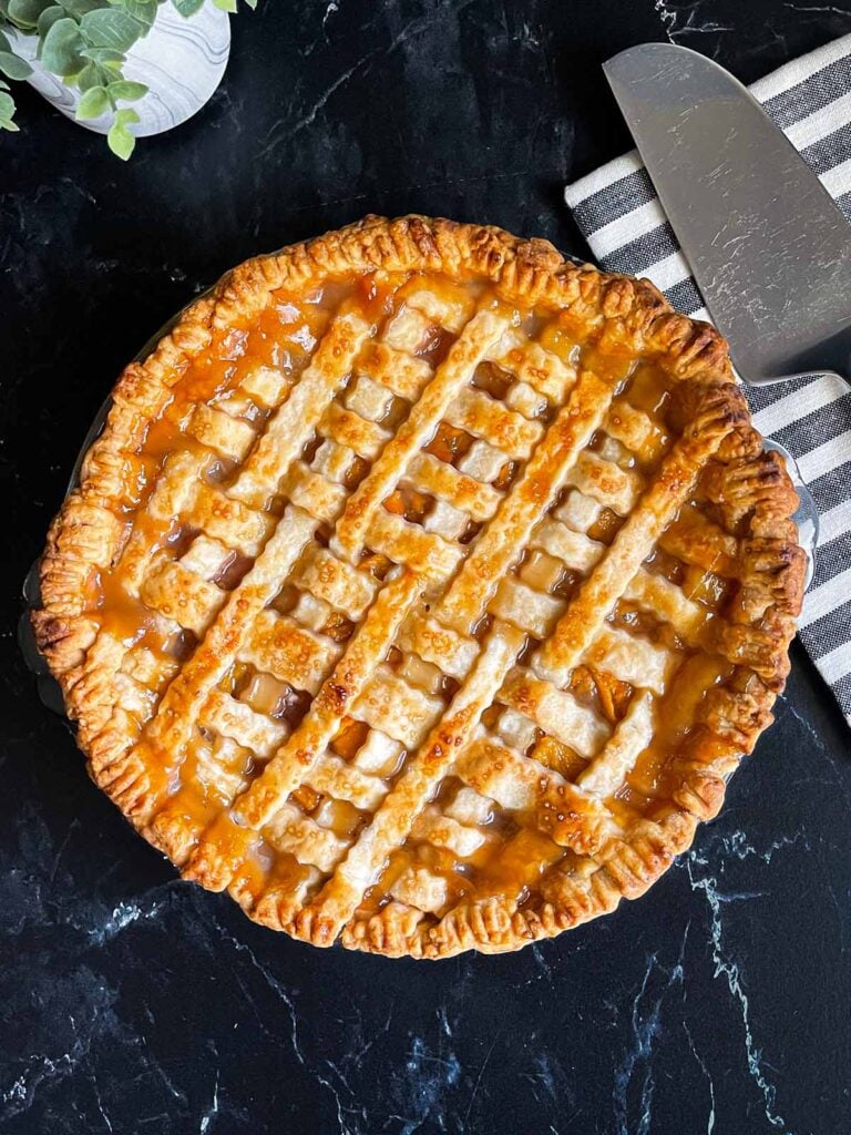 A peach pie with a lattice crust with a striped napkin and pie slicer in the background.