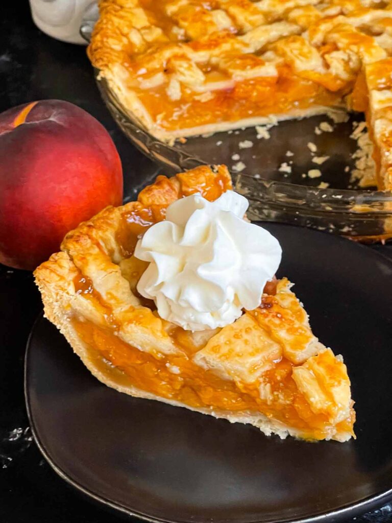 A slice of peach pie with whipped cream on a dark plate.