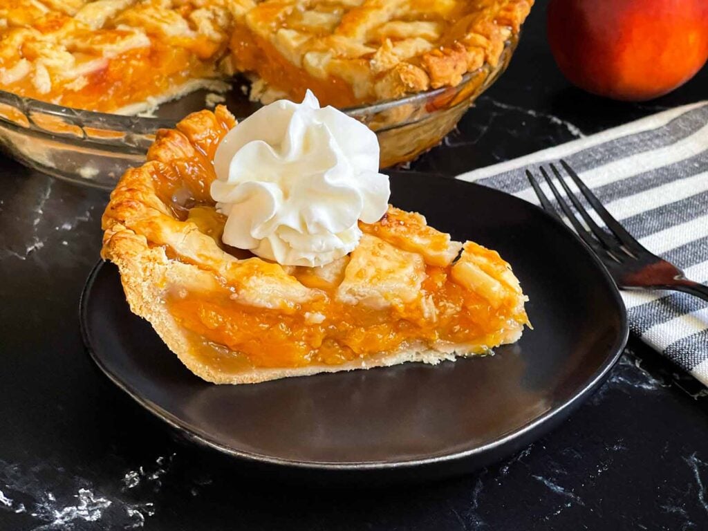 A slice of peach pie with whipped cream on a dark plate.